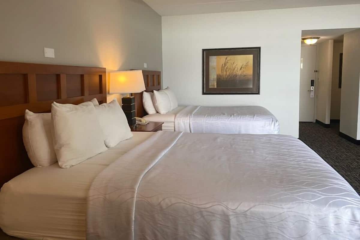 A hotel room with two beds, a nightstand with a lamp, a framed picture on the wall, and a door leading to another room.