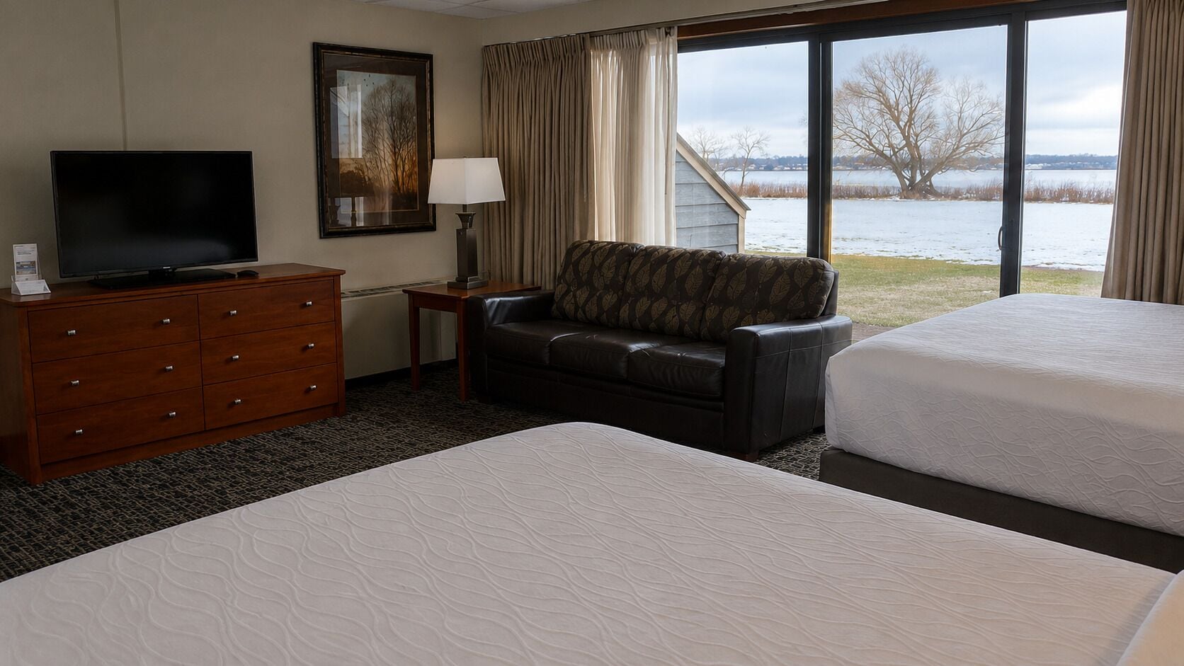 Habitación de hotel con una cama grande, un sofá, un televisor, un aparador y una ventana con vista al lago.