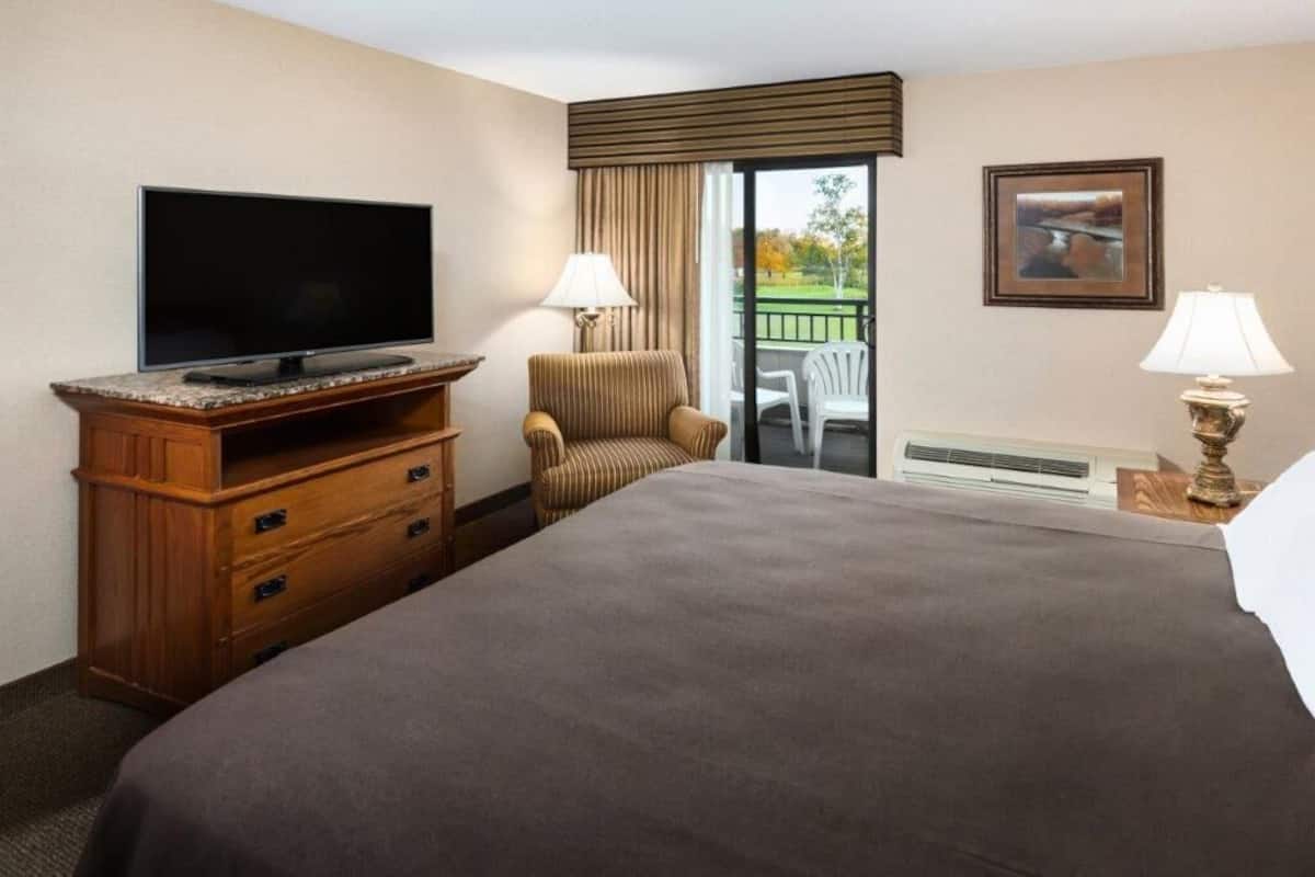A hotel room with a large bed, a flat-screen TV, a wooden dresser, a chair, a balcony with a view, and a framed picture on the wall.
