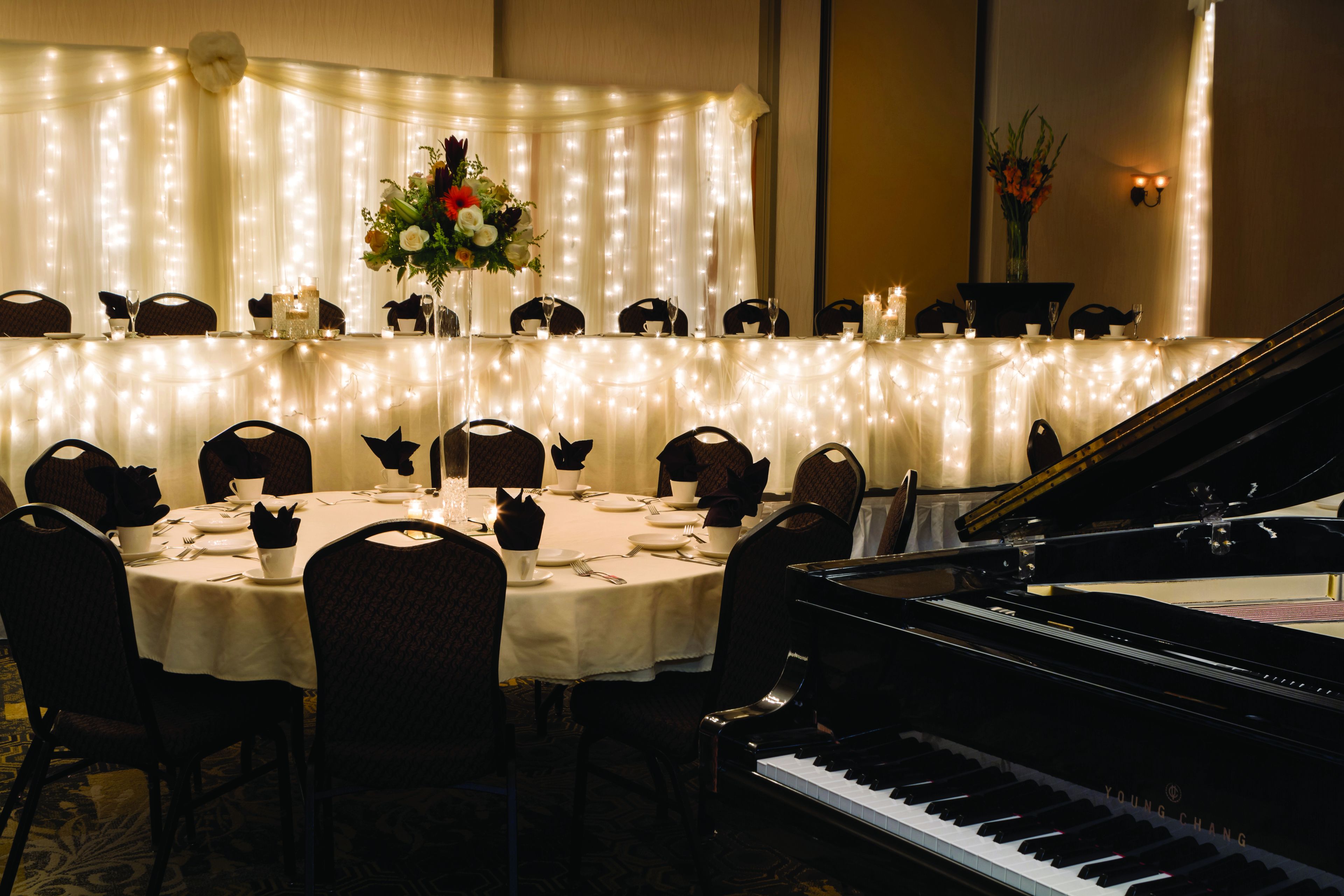A banquet hall with a piano, round tables, chairs, and string lights.
