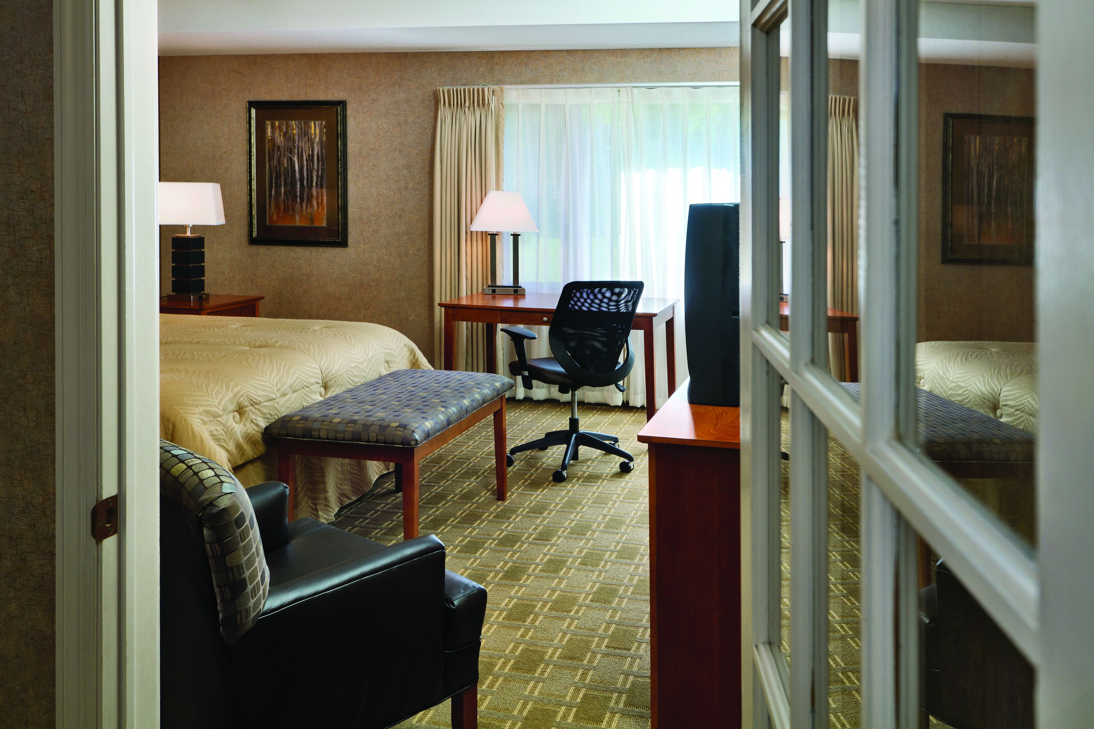 A hotel room with a bed, a desk with a chair, a small table with a lamp, and a window with curtains.