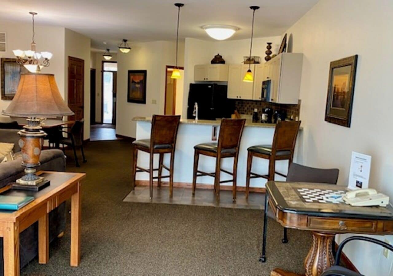 Un área de comedor con una mesa, sillas y una lámpara. Se observa una cocina al fondo, con armarios y un refrigerador.