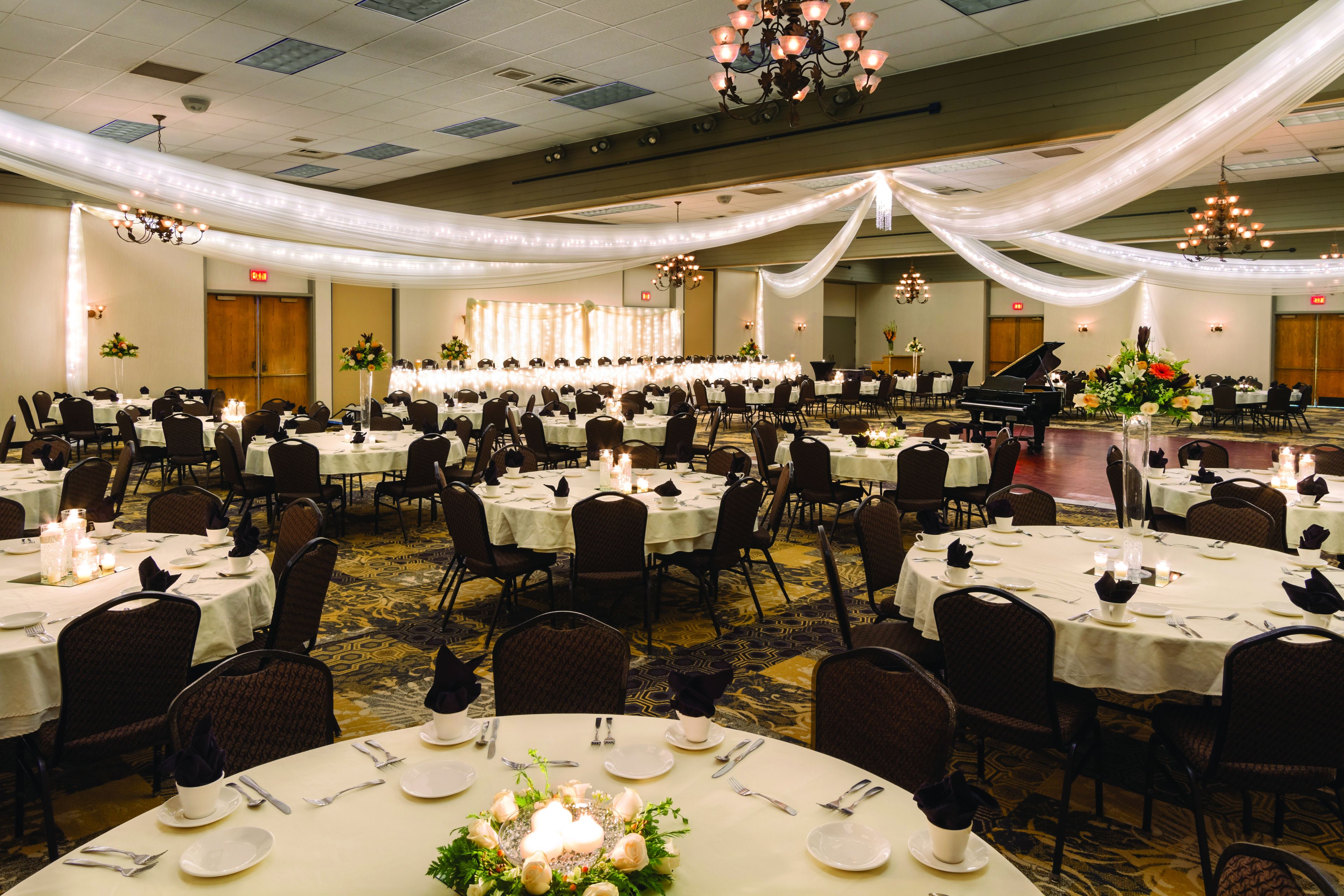 A banquet hall with round tables set for a formal event, featuring chandeliers and draped white fabric.