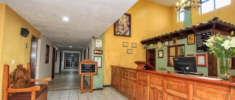 A hotel lobby with a reception desk, wooden benches, a chandelier, and framed pictures on the walls.