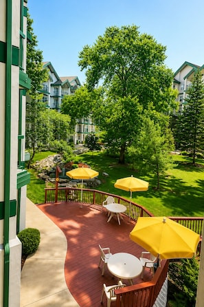 A deck with outdoor furniture and umbrellas, surrounded by greenery and a building. - Carriage Place (Branson)