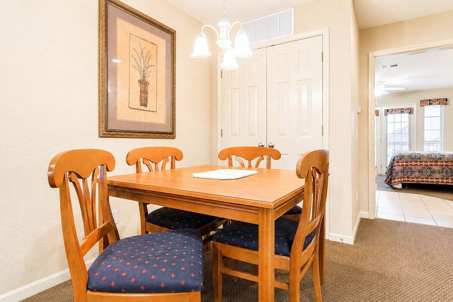 A dining area with a wooden table and four chairs, a framed picture on the wall, and a door leading to another room.