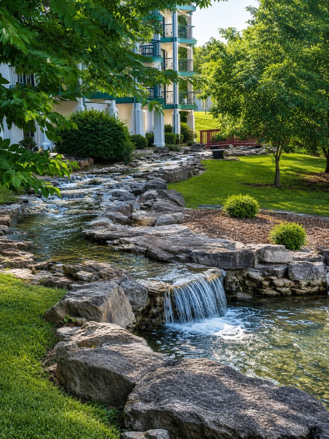 A landscaped garden with a waterfall, a building with balconies, and a well-maintained lawn.