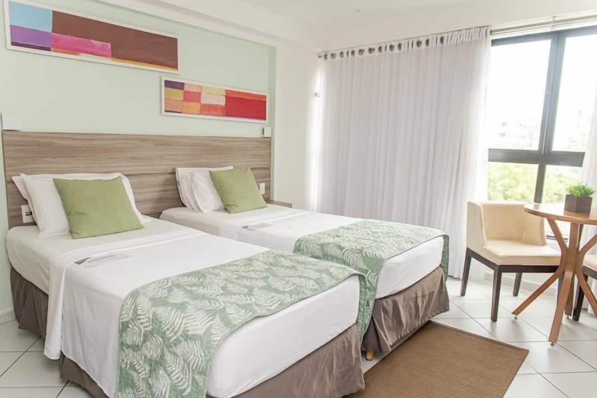 Habitación de hotel con dos camas, una mesita, una silla y una ventana con cortinas.