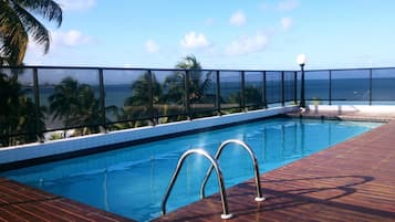 A pool with a wooden deck and metal railings, overlooking a beach with palm trees and the ocean.