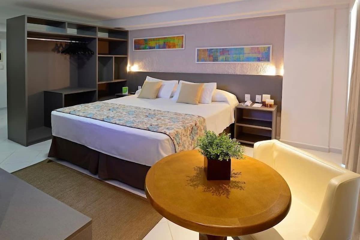 Habitación de hotel con cama, mesita de noche con planta, silla, armario y una obra de arte en la pared.