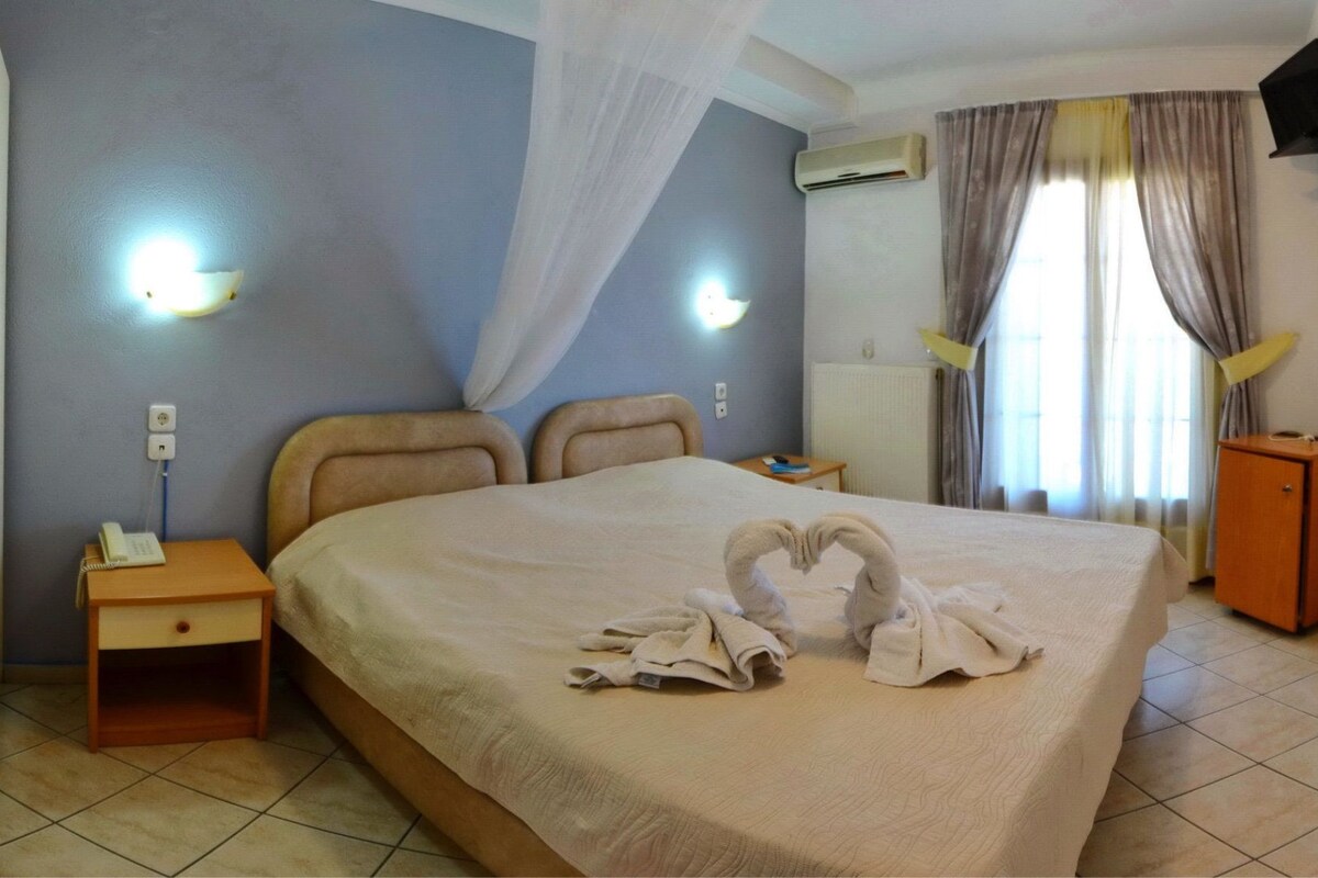 A hotel room with a bed, a bedside table with a phone, an air conditioning unit, a small cabinet, and curtains.