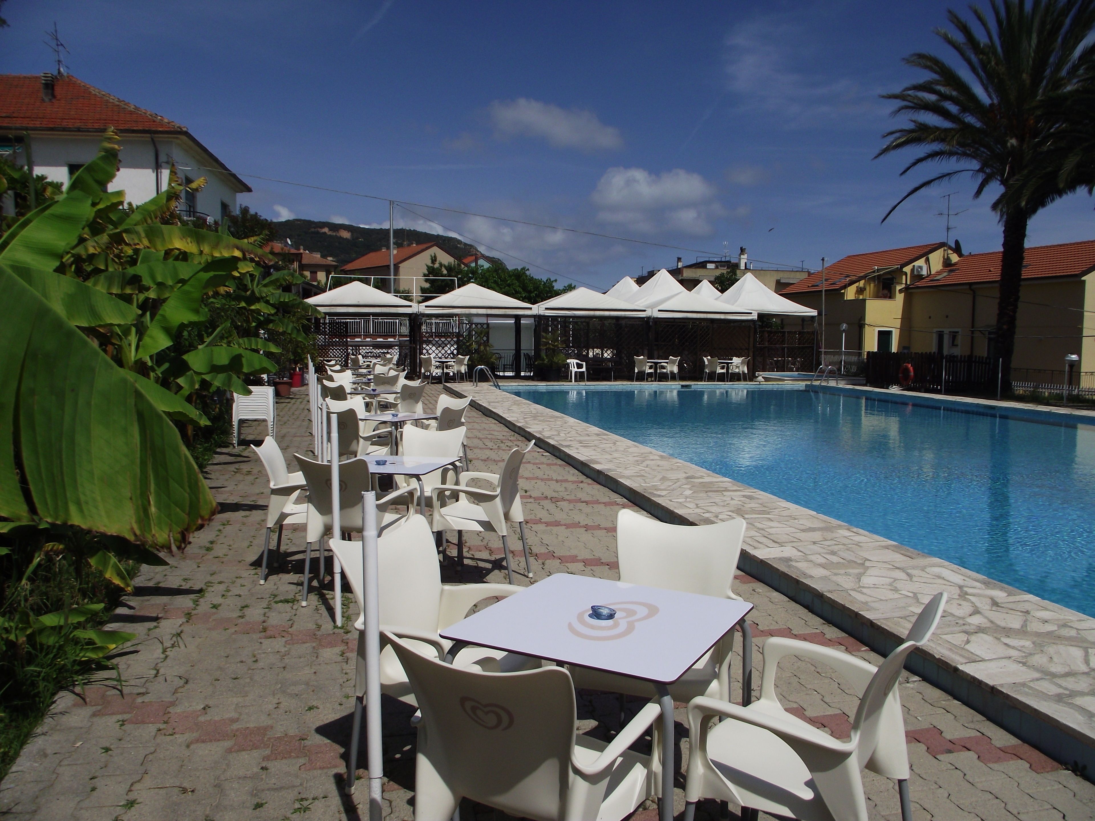 A poolside area with white plastic chairs and tables, a swimming pool, and umbrellas.