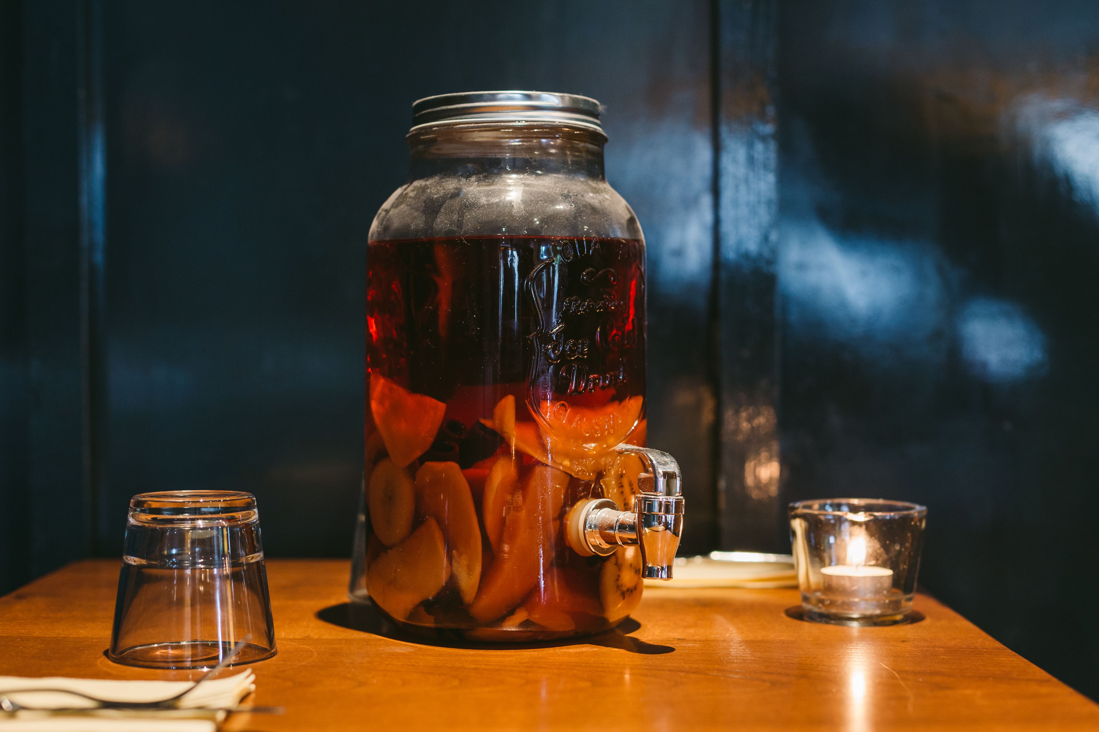 A large glass jar with a tap, filled with a dark liquid and slices of fruit, placed on a wooden table with two small glasses and a candle.