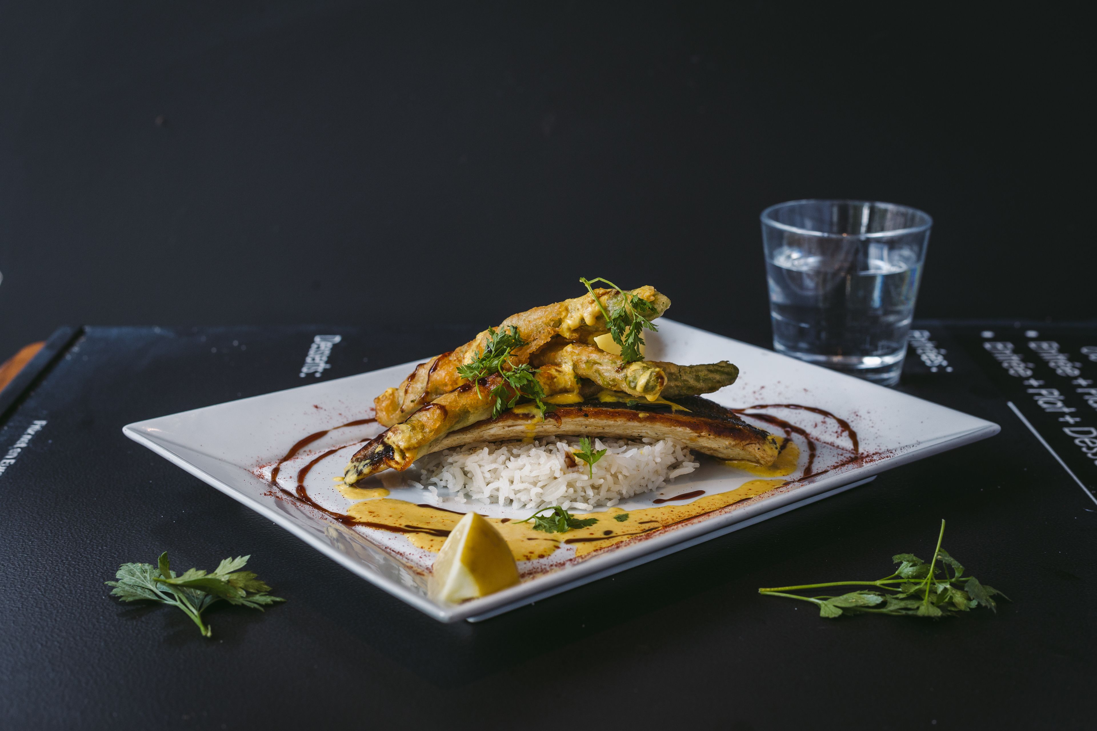 A square plate with rice, grilled fish, and lemon on a black table.