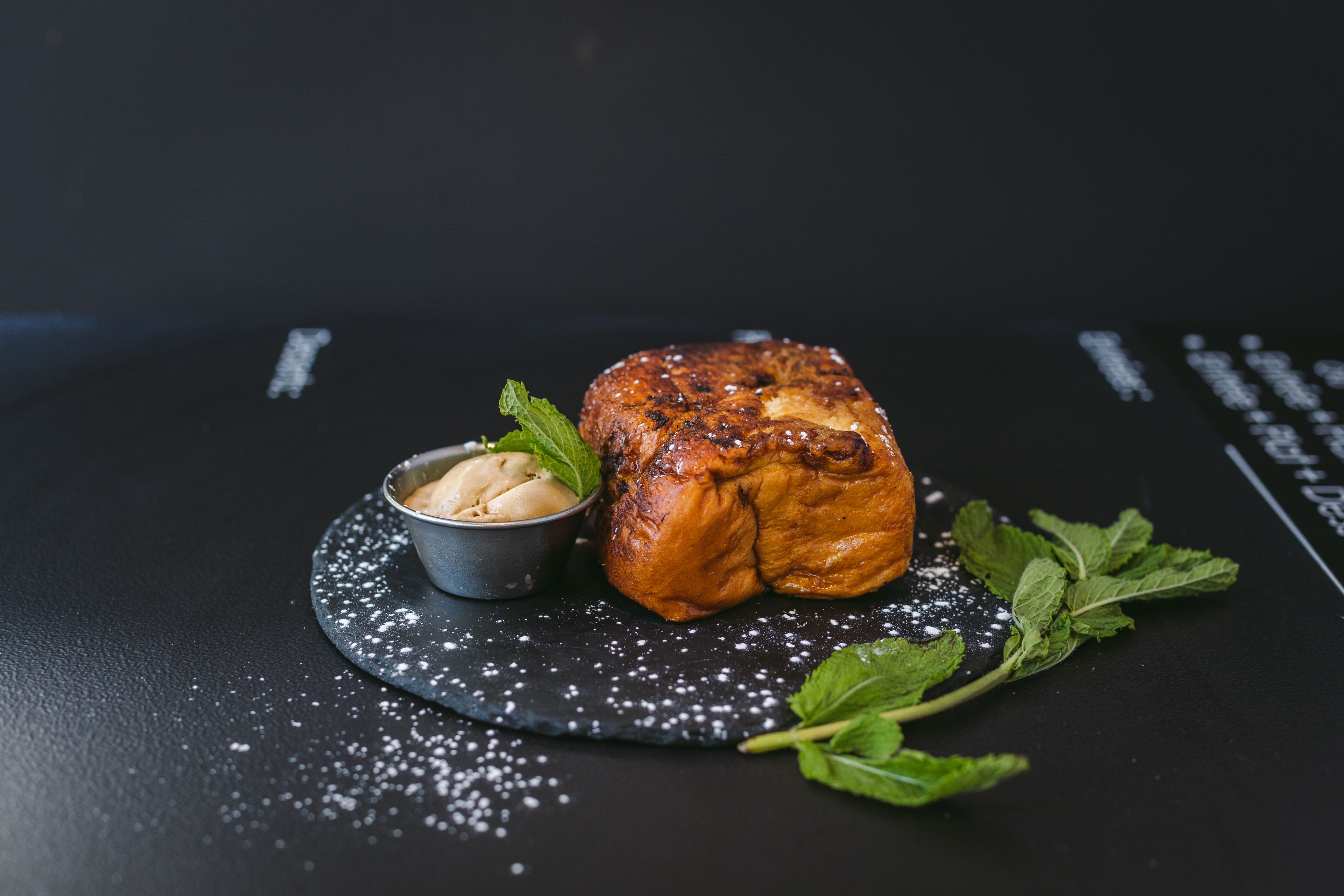 A loaf of bread with a dusting of white powder, a small bowl of butter, and fresh mint leaves on a dark surface.