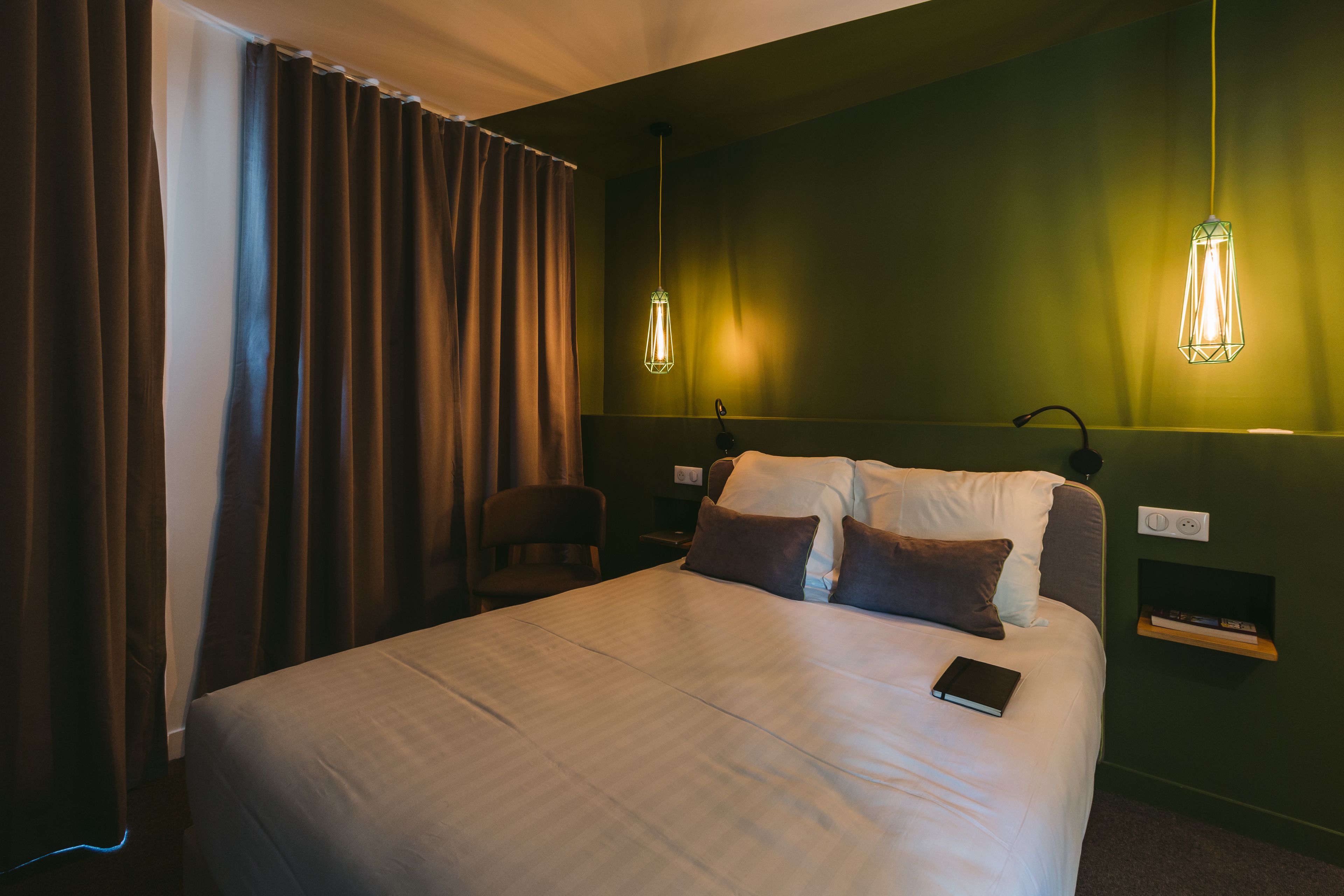 Een net opgemaakt bed met kussens en een boek, een stoel, een nachtkastje met een lamp, en een wandplank.