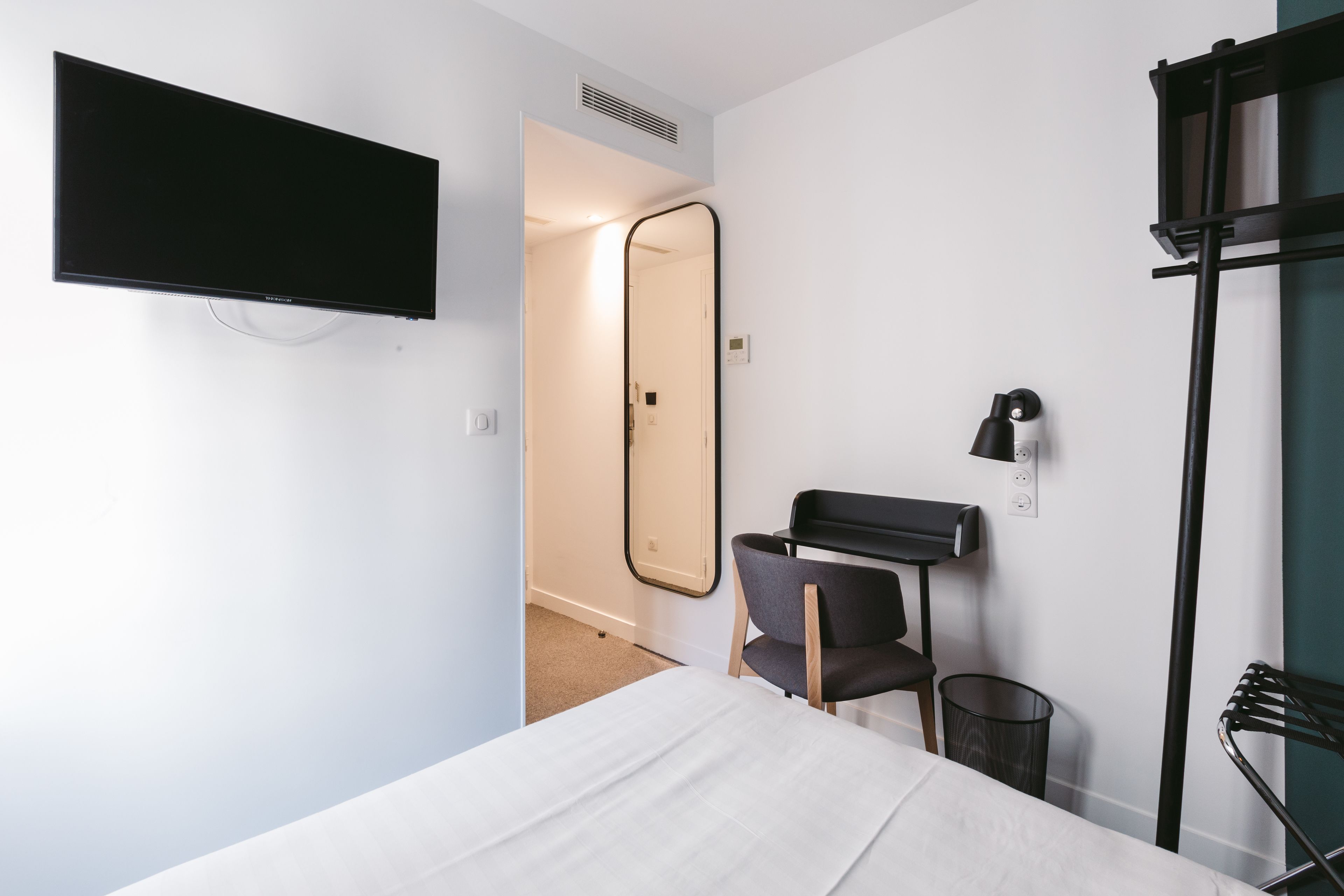 A hotel room with a bed, a desk, a chair, a TV, a mirror, and a trash bin.