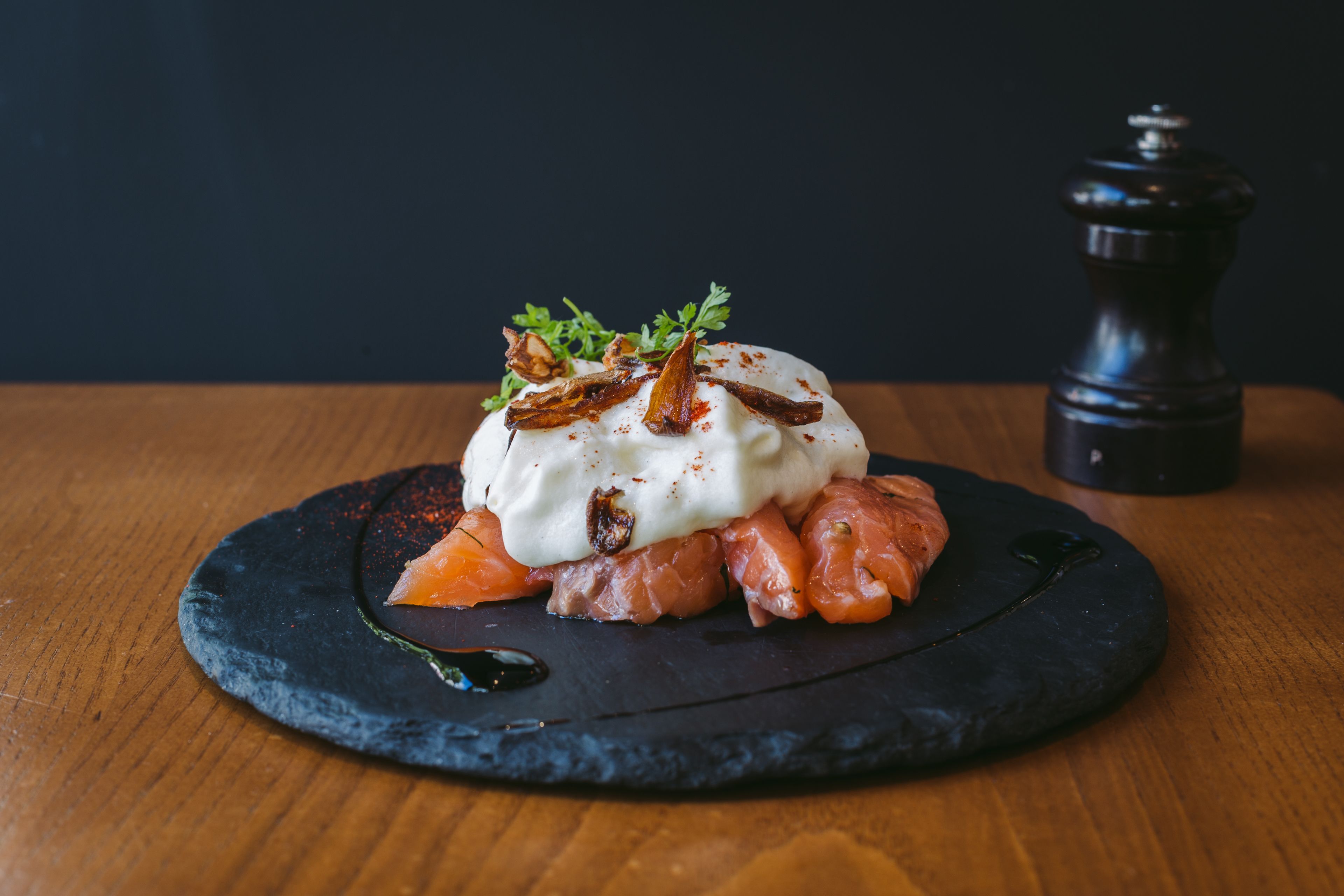 Un plato con cobertura cremosa, adornado con hierbas y elementos fritos, sobre una losa de pizarra oscura.