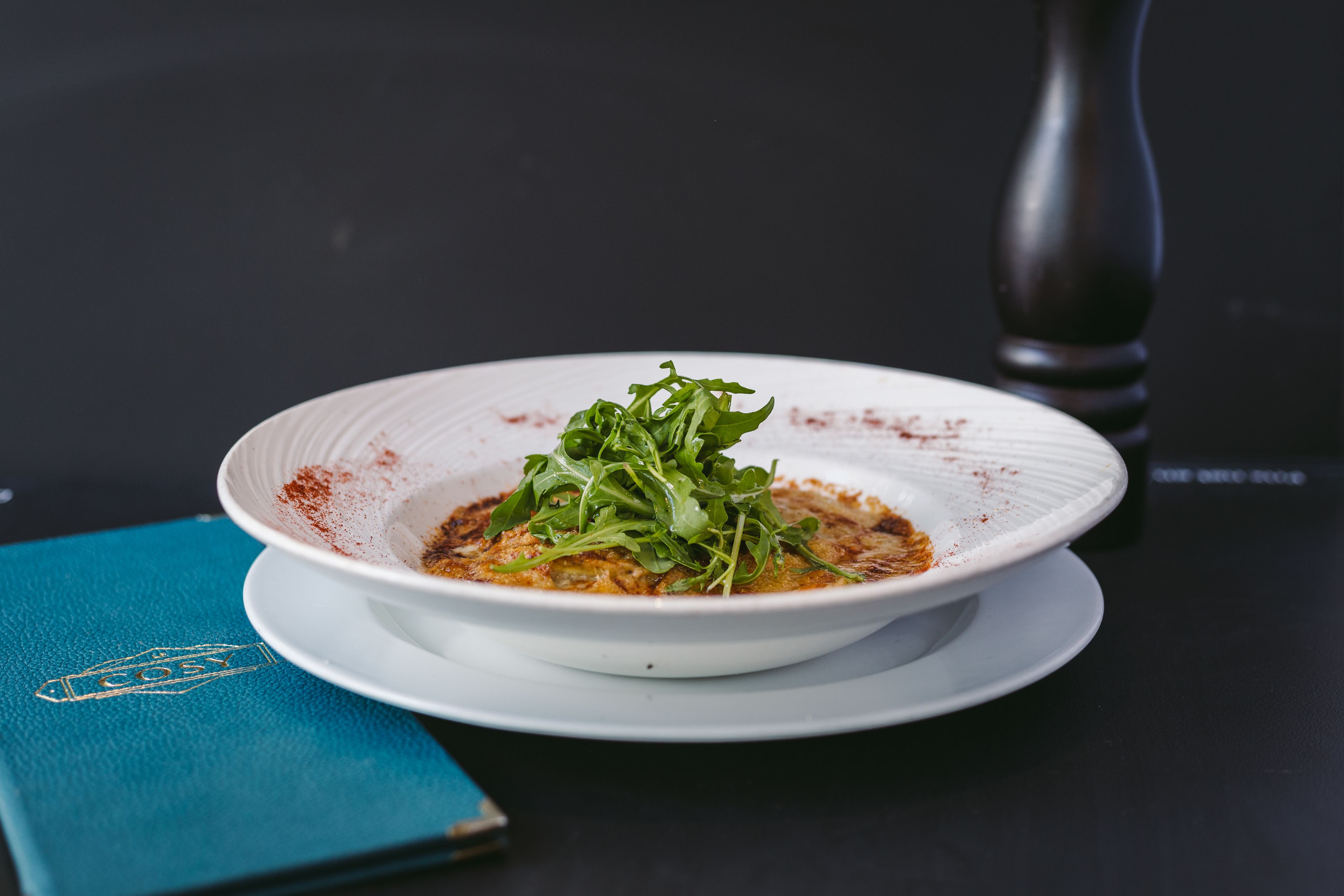 A white bowl of food with greens and spices on a dark table, a salt shaker, and a blue menu.