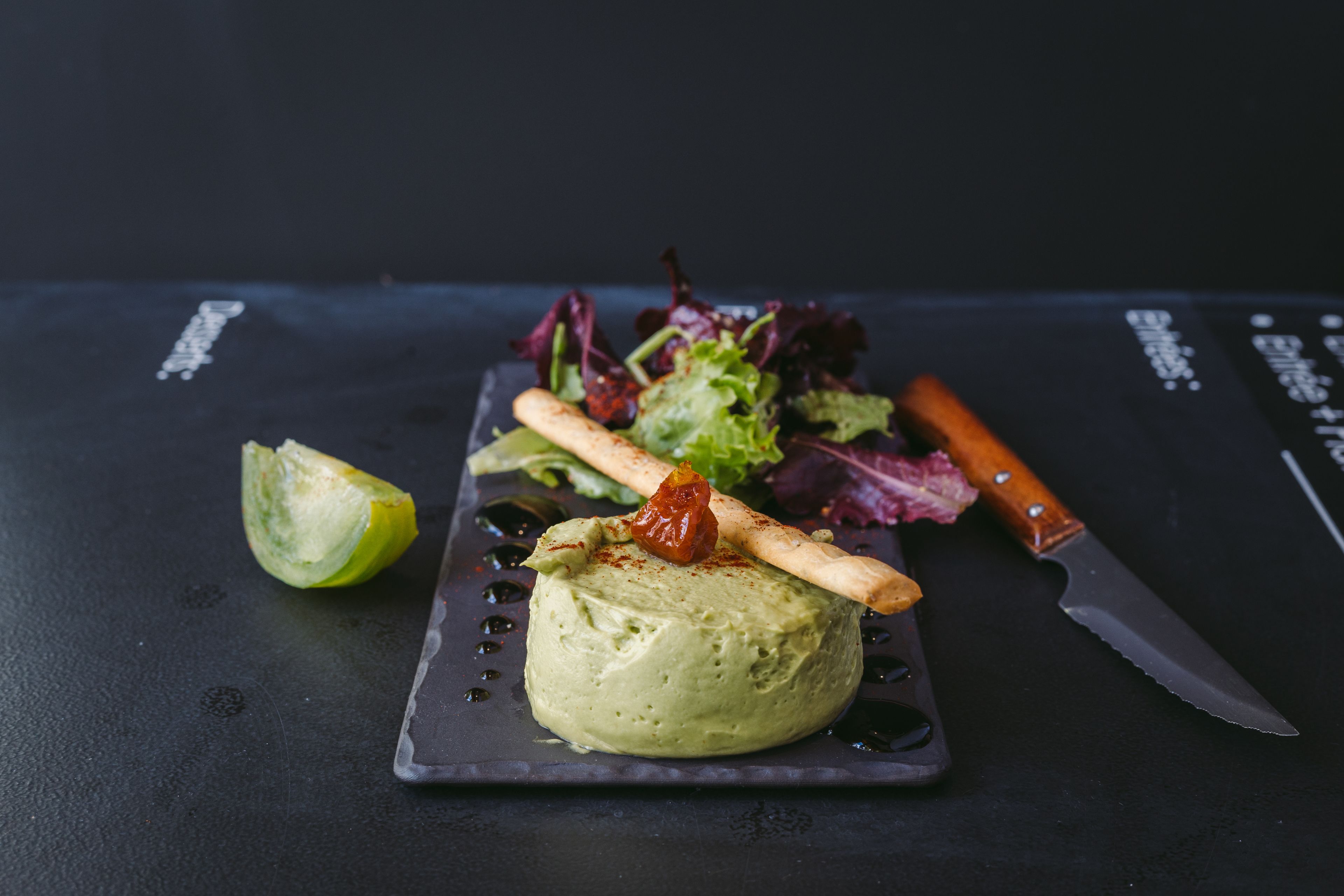 A plate with a green spread, a wooden skewer, a lime half, and a knife on a dark surface.