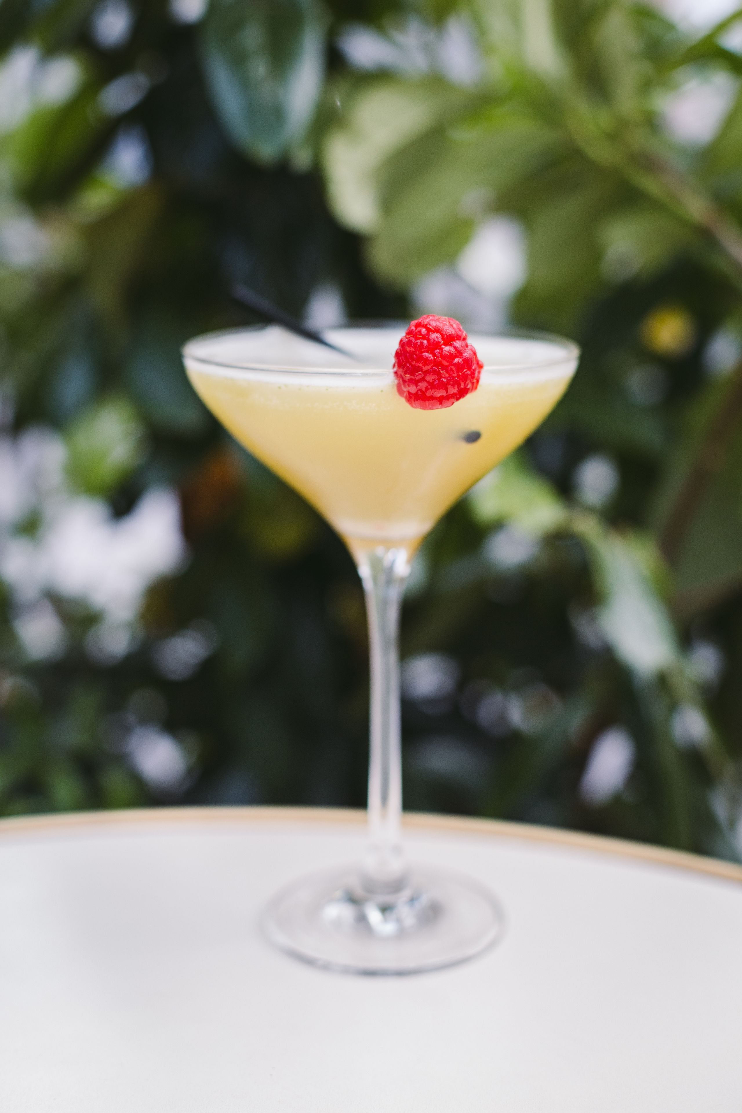A cocktail in a martini glass with a raspberry garnish, set against a blurred green background.