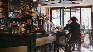 Zona de bar con un barman, estantes con botellas y vistas a la calle a través de amplios ventanales.