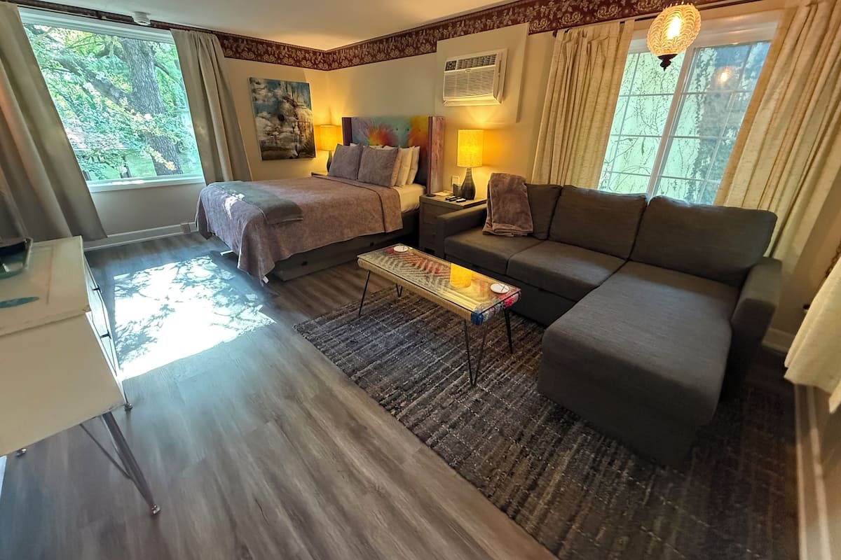 A bedroom with a bed, a sofa, a small table, and a nightstand. There is a window with curtains, a painting on the wall, and a lamp on the nightstand.