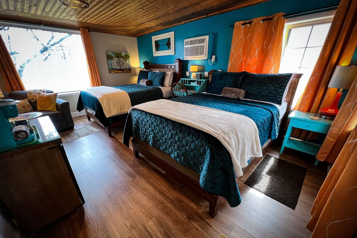 A room with two beds, a wooden ceiling, a window with curtains, and a desk with a lamp.
