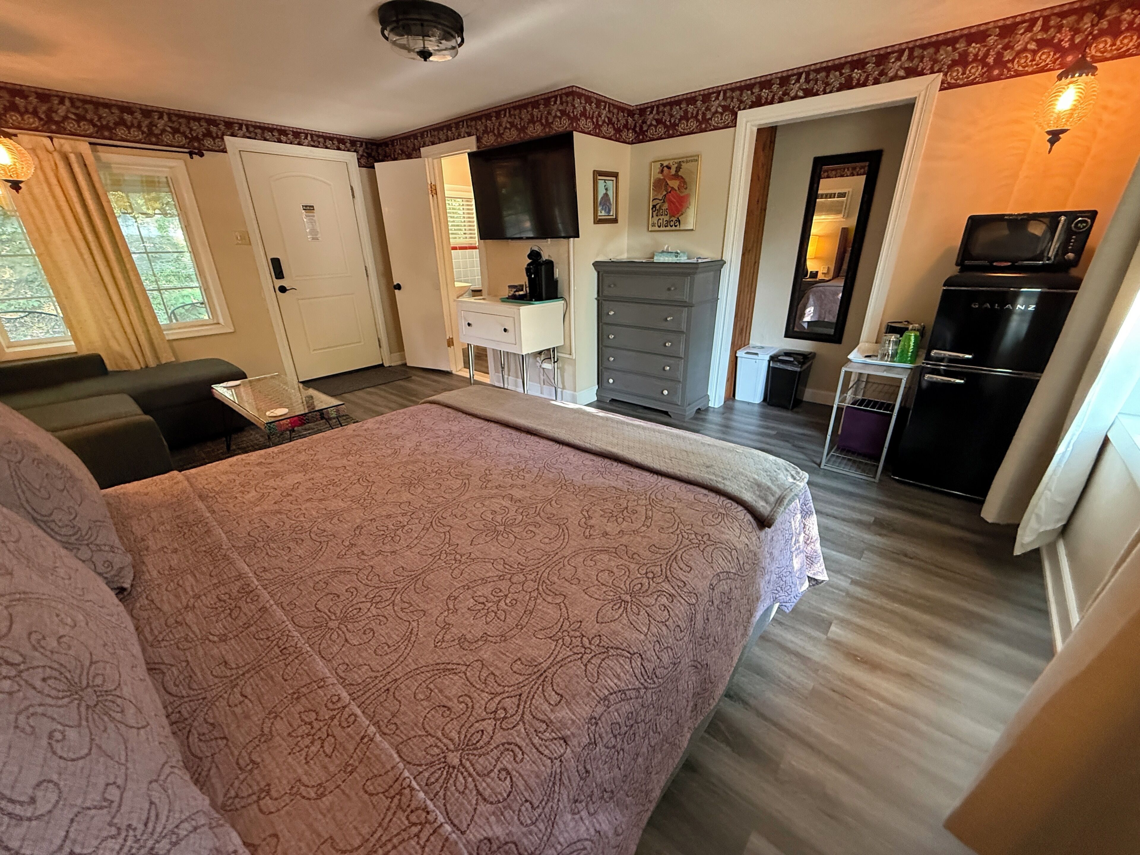 A bedroom with a large bed, a desk, a dresser, a television, and a kitchenette.