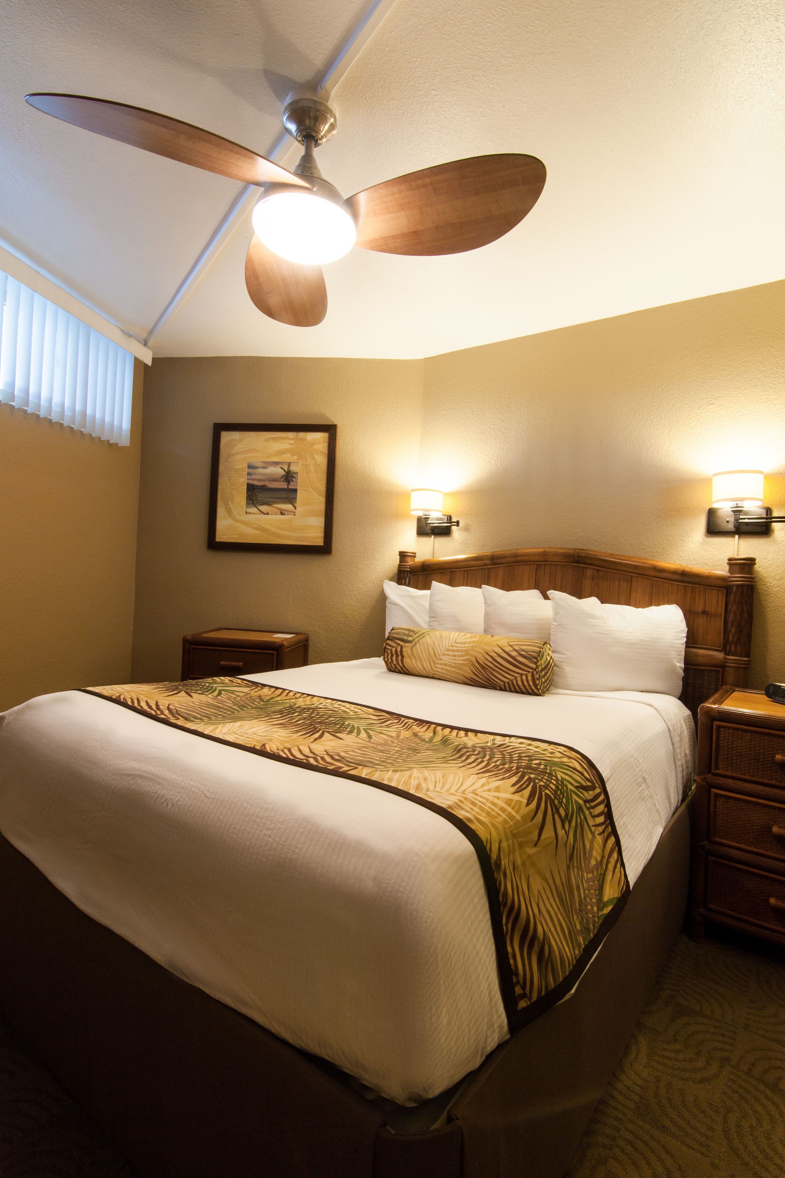 A hotel room with a bed, a ceiling fan, a nightstand, and a wall-mounted light fixture.