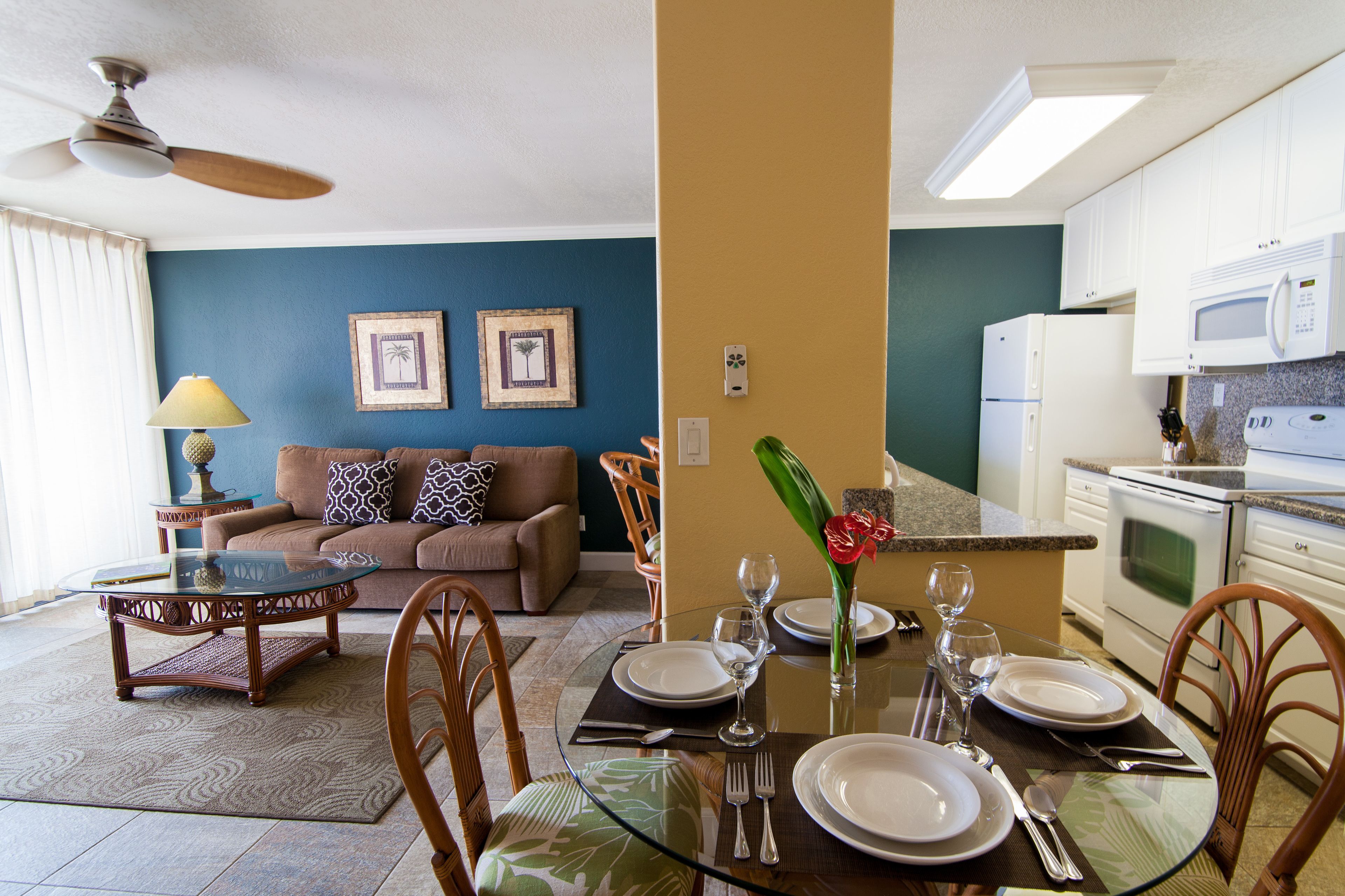 A dining area with a glass table set for four, a sofa, and a kitchen in the background.