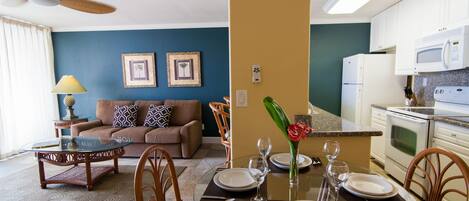 A dining area with a glass table set for four, a sofa, and a kitchen in the background.