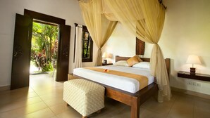 A spacious bedroom with a large bed, a bedside table with a lamp, and a view of a tropical garden through an open door.
