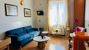 A modern living room with a blue sofa, a round coffee table, and a small side table. There is a window with sheer curtains, a potted plant, and a decorative lamp. - Bon Ton Design Rooms by DOT Suites (Rome)