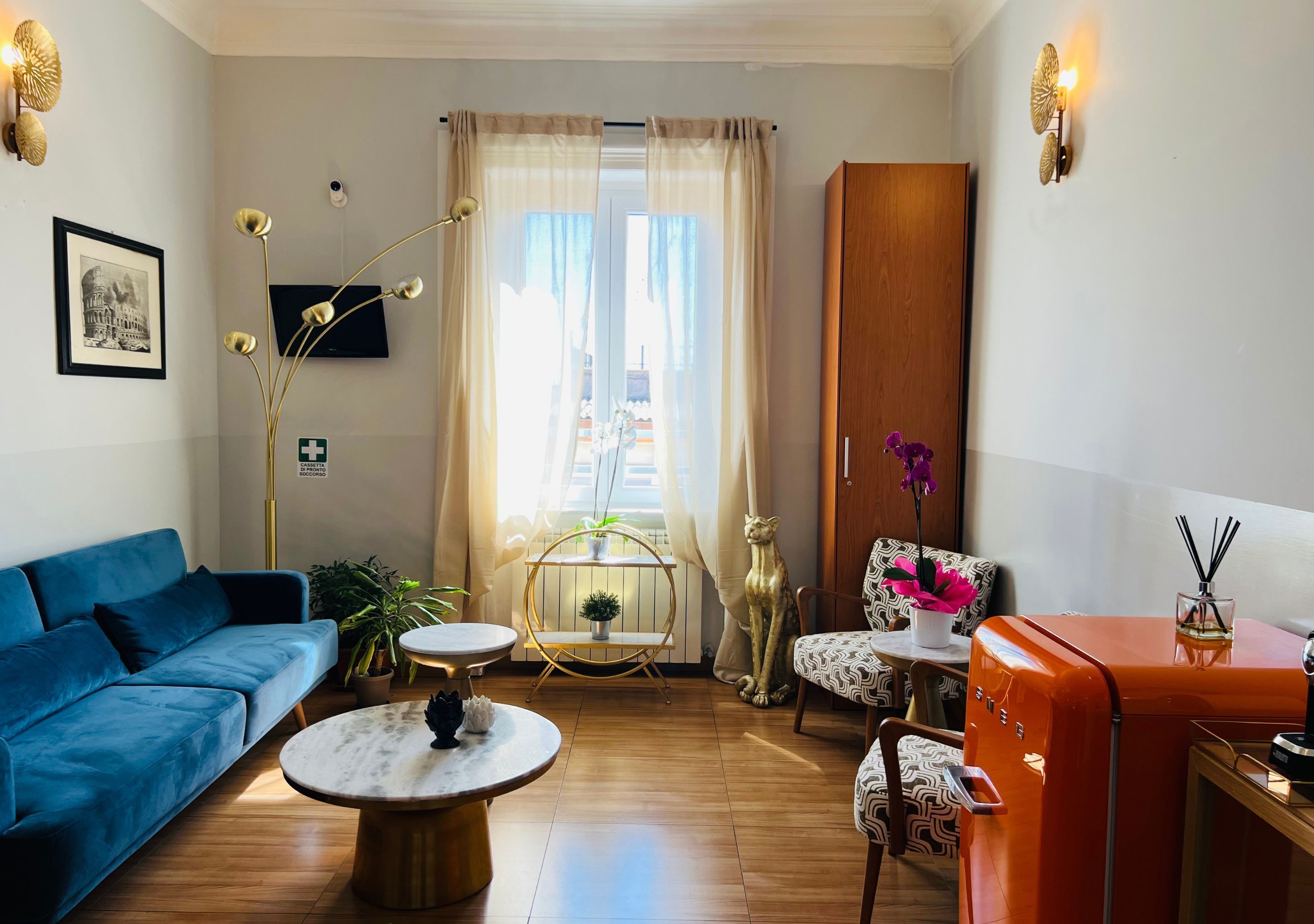 Ein Wohnzimmer mit blauem Sofa, rundem Couchtisch, einem kleinen Tisch mit Pflanze, einer hohen Stehlampe, einem Schrank und einem Fernseher an der Wand.