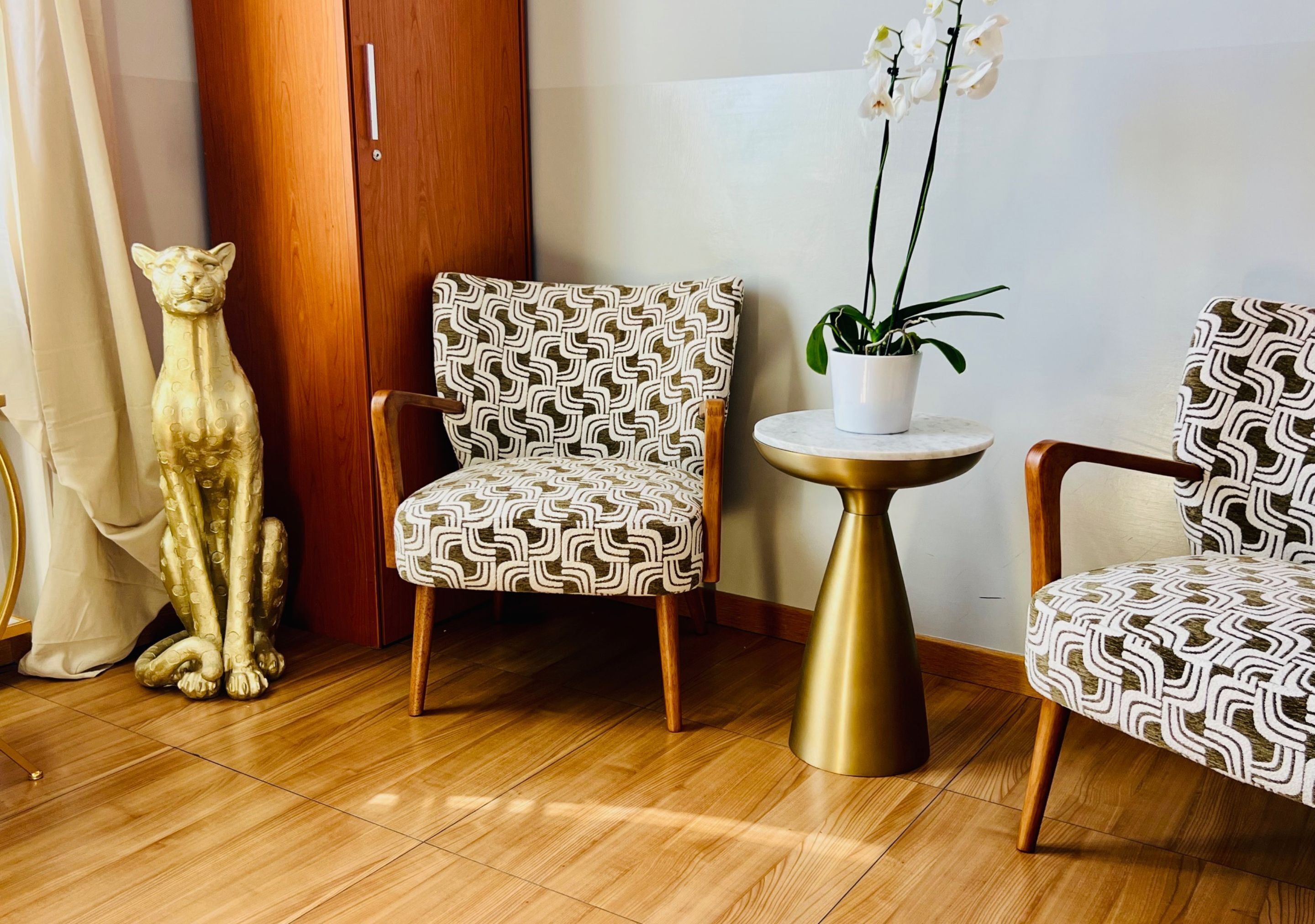 A room with a wooden floor, a patterned armchair, a small round table with a plant, and a decorative cat statue.