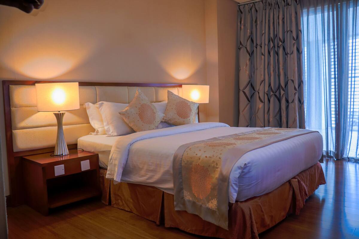 Ein Hotelzimmer mit einem großen Bett, zwei Nachttischlampen, einem Nachttisch und einem Fenster mit Vorhängen.