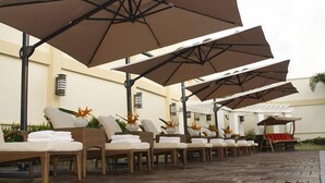 Une rangée de chaises longues dotées de coussins blancs et de parasols bruns, disposées sur une terrasse dallée.