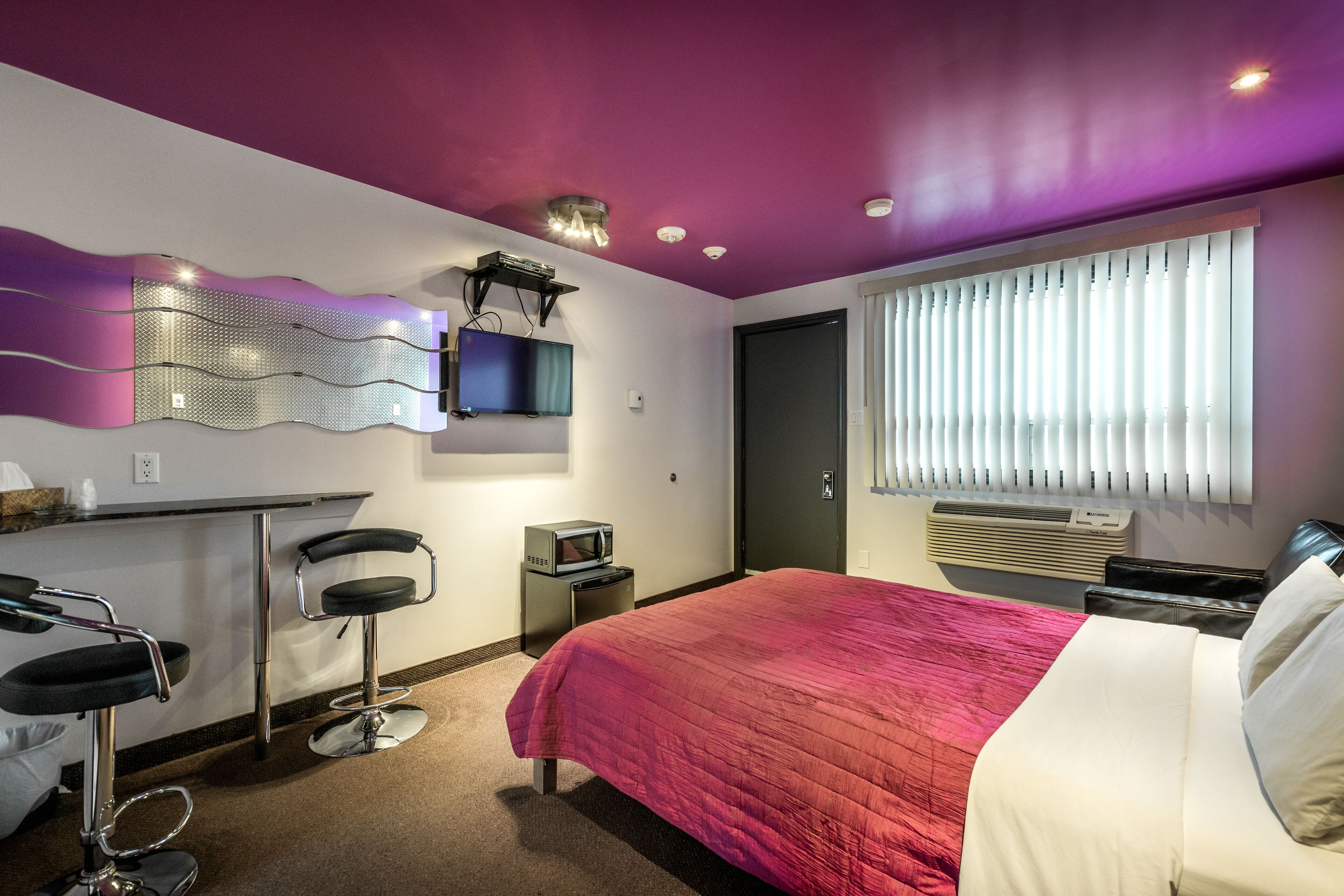 Habitación de hotel con cama, escritorio con silla, televisor y ventana con persianas.