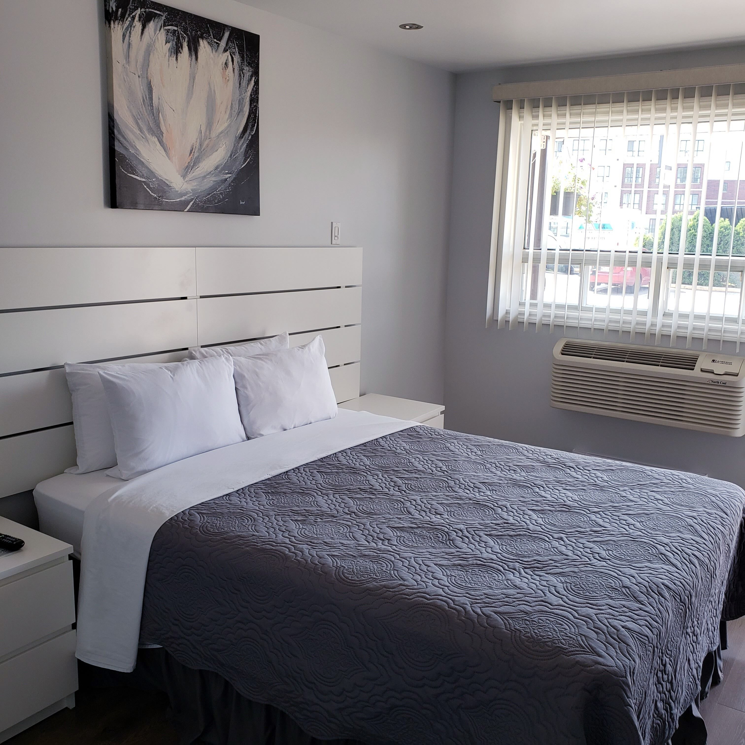 Un dormitorio moderno con una cama, mesitas de noche blancas, un cuadro abstracto grande y una ventana con persianas.