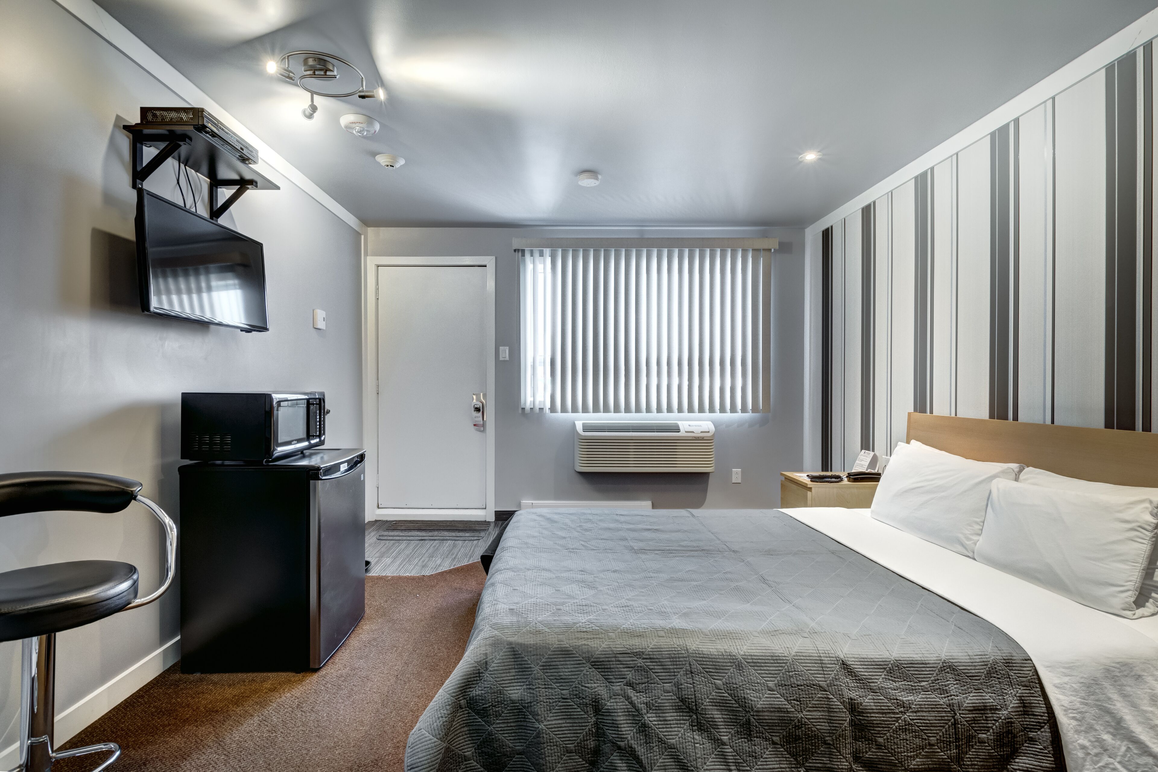 A modern hotel room with a bed, a desk with a microwave, a chair, a window with blinds, and a ceiling light fixture.