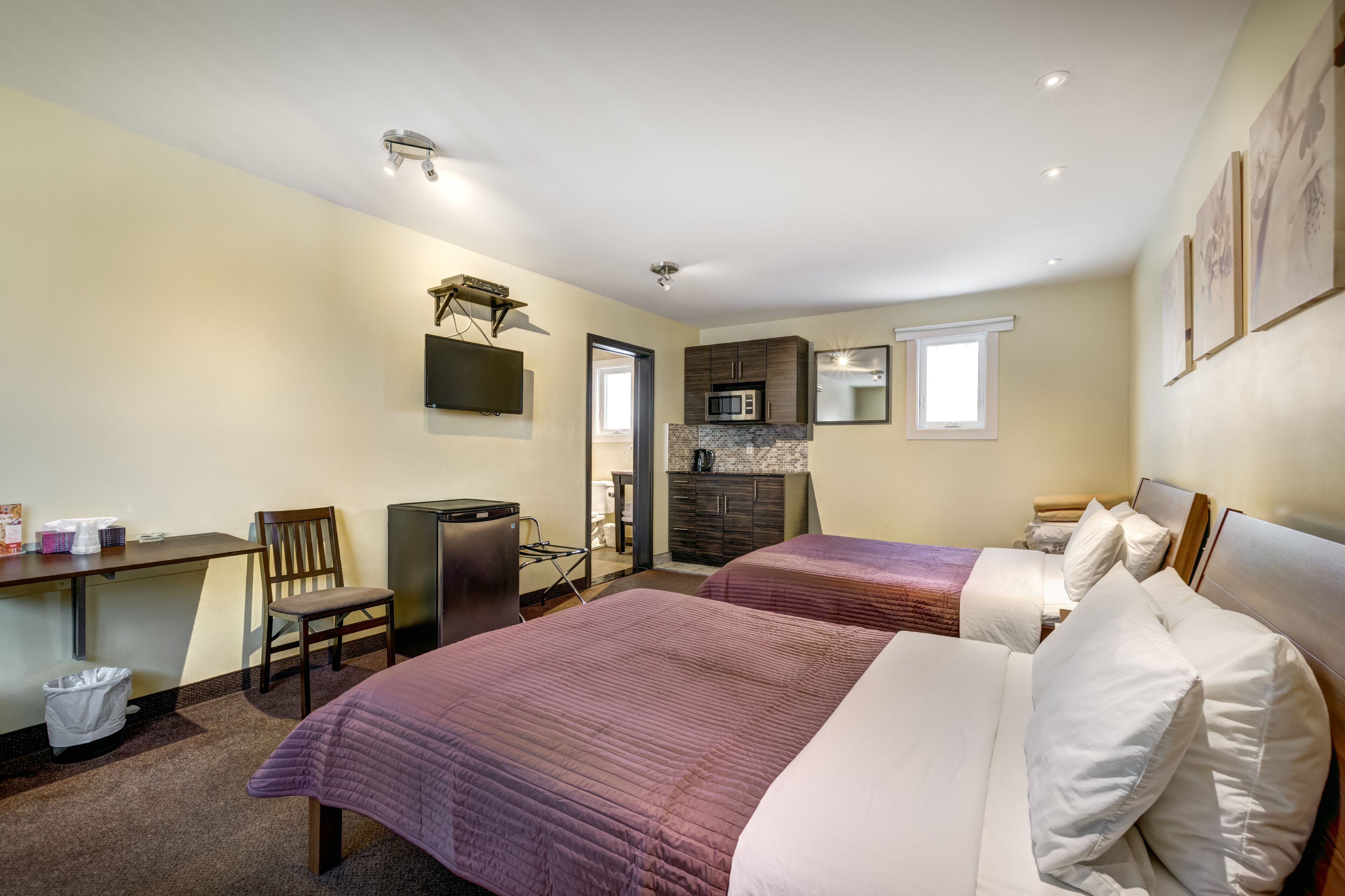 A hotel room with two beds, a desk, a chair, a TV, a microwave, and a small kitchenette.