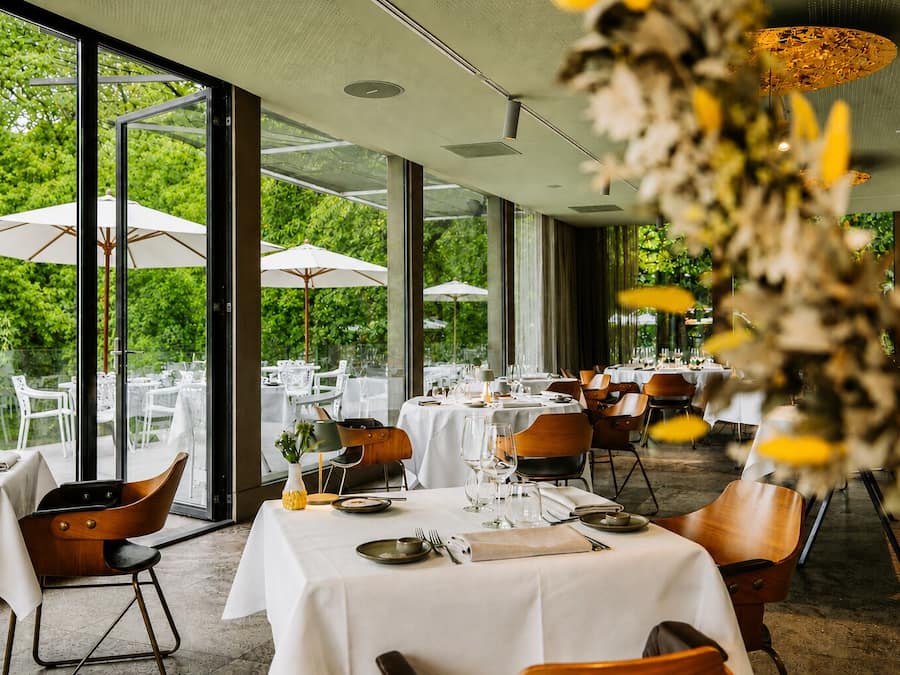 An outdoor dining area with tables set for two, surrounded by glass walls, and a view of greenery outside.