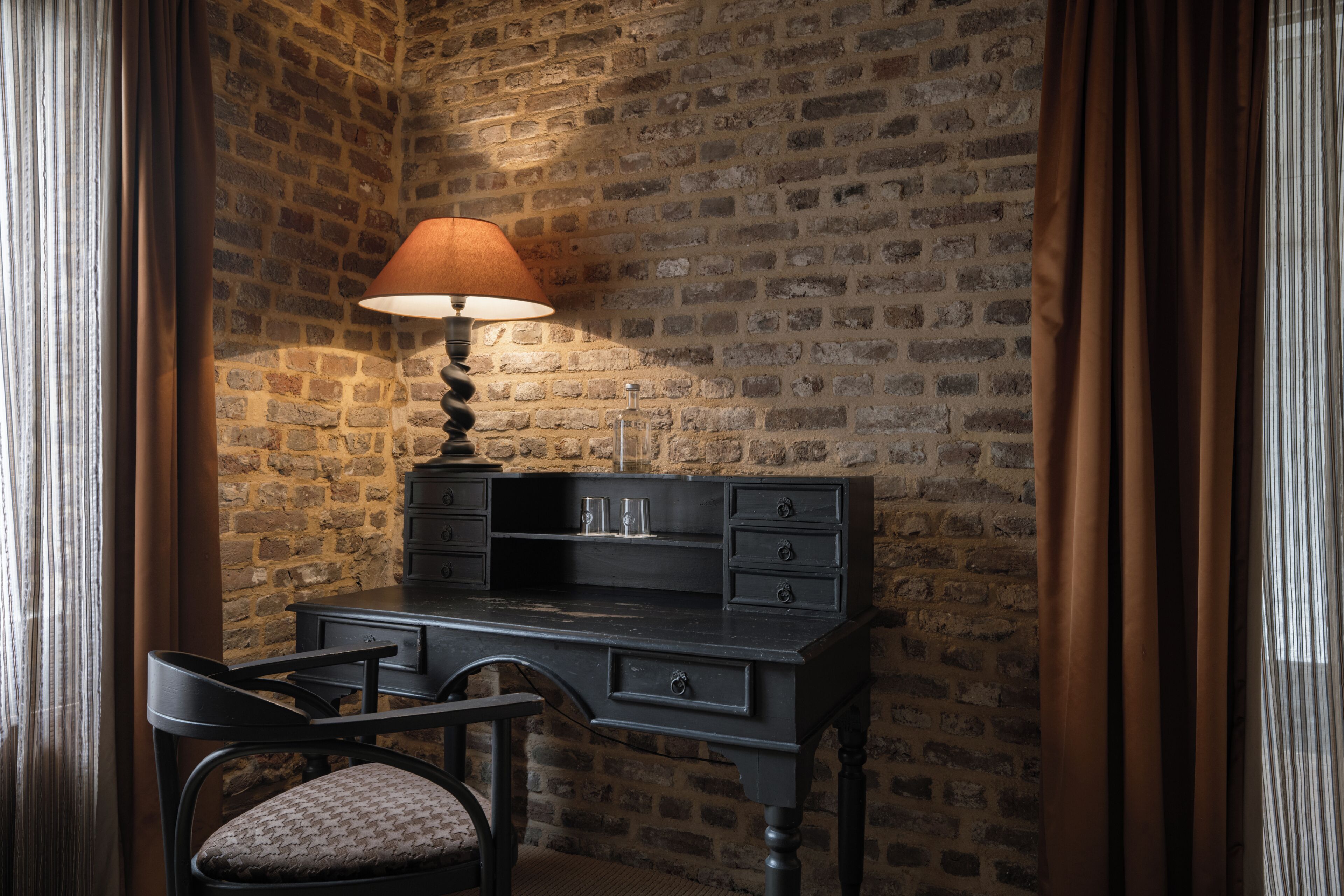 Una habitación de esquina con pared de ladrillo, un escritorio con cajones, una silla y una lámpara.