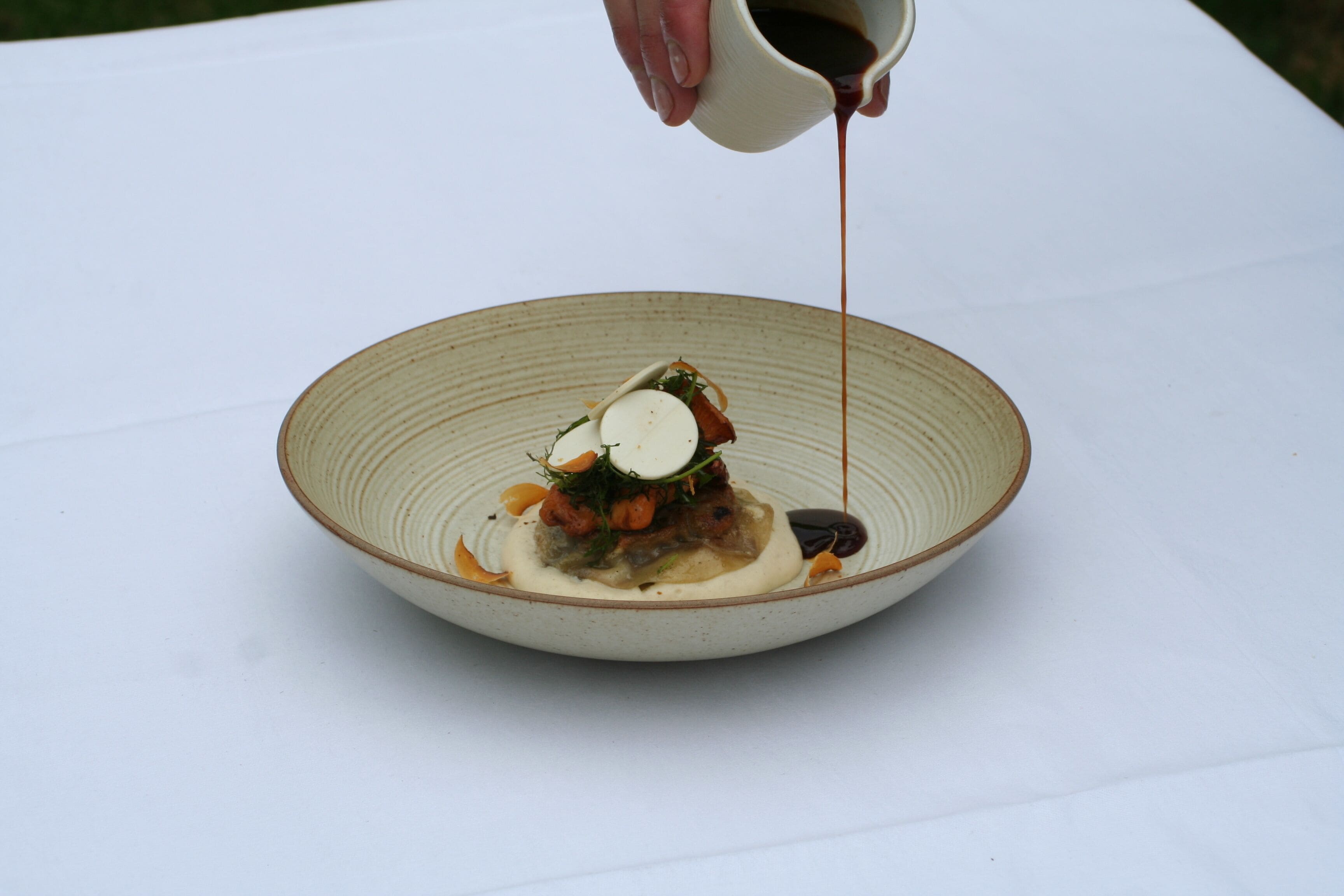 A white table with a ceramic bowl containing a dish with various food items and sauce being poured.