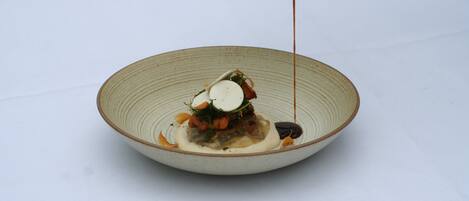 A white table with a ceramic bowl containing a dish with various food items and sauce being poured.