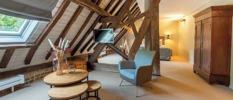 A cozy attic room with a wooden ceiling, a television mounted on the wall, a round table with two chairs, and a cabinet.