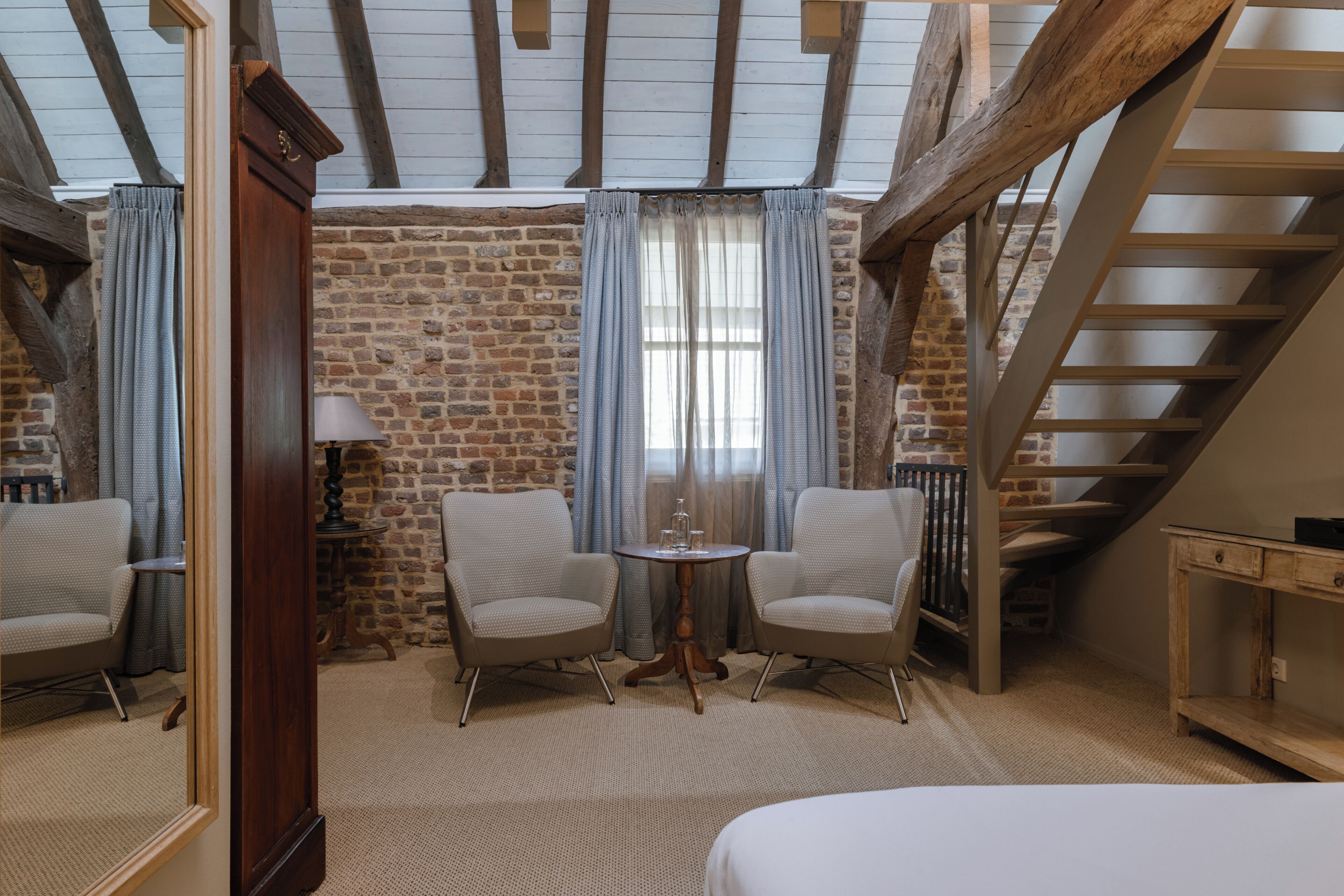 A cozy room with a brick wall, a wooden ceiling, a staircase, two chairs, a small table, and a bed.