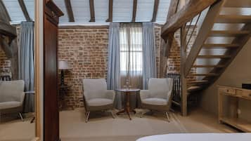 A cozy room with a brick wall, a wooden ceiling, a staircase, two chairs, a small table, and a bed.