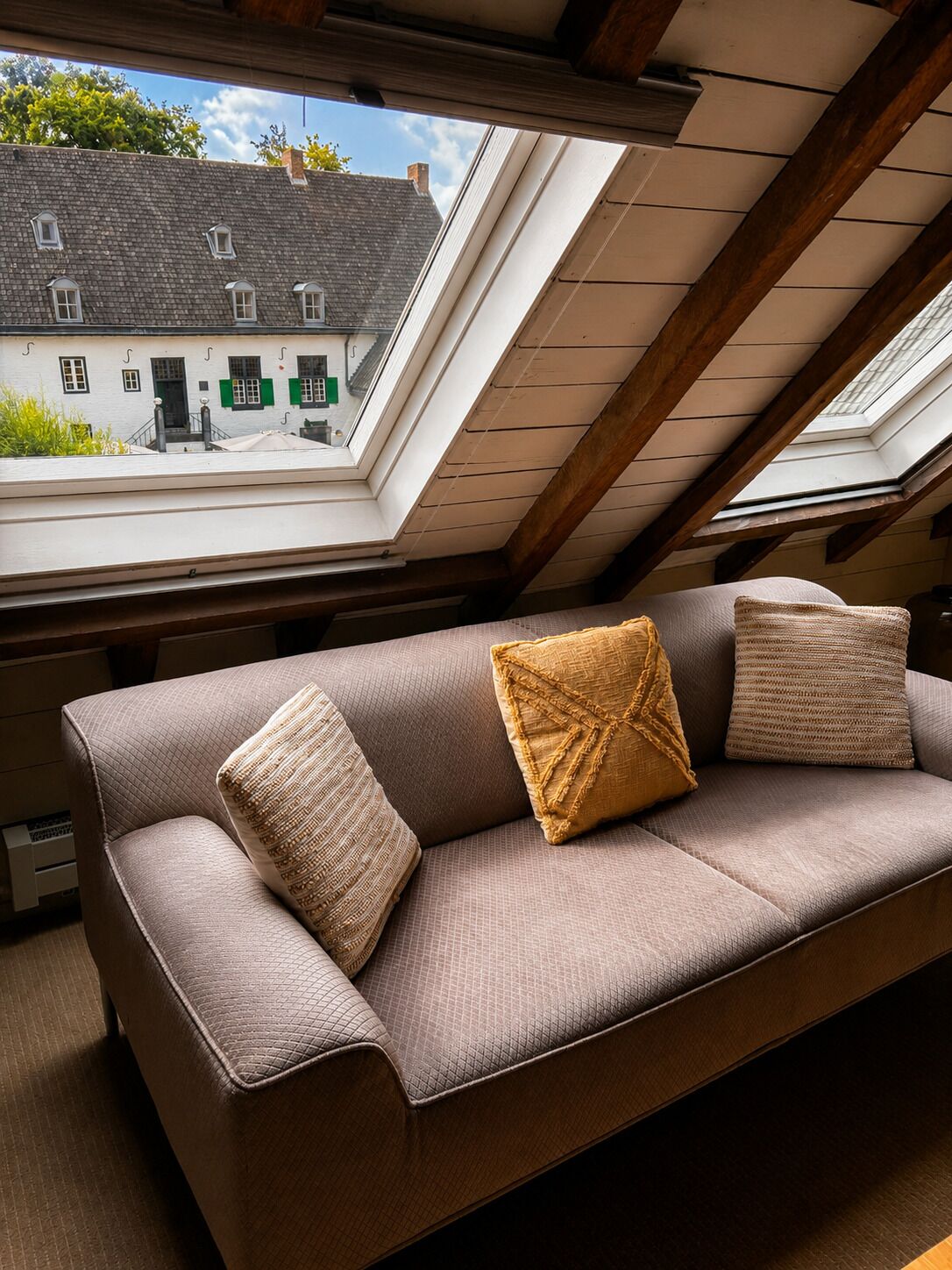 Una habitación con techo inclinado, un sofá marrón con cojines y vistas a un edificio a través de un tragaluz.